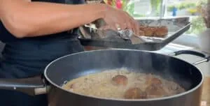 Person cooking creamy mushroom meatballs in a skillet, placing fresh meatballs onto a tray in a bright kitchen.
