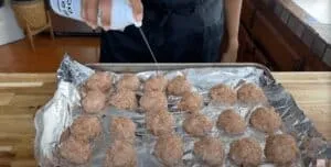 Person spraying oil on meatballs on a foil-lined baking sheet, preparing for cooking in a kitchen.