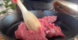 Cooking ground meat in a skillet with a wooden spoon in a kitchen setting.