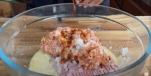 Mixing ingredients in a glass bowl for a meatloaf recipe with seasoning and sauce being added.