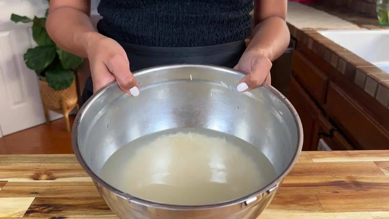 Person holding metal bowl with rice soaking in water on a wooden countertop, preparing for cooking in kitchen setting.