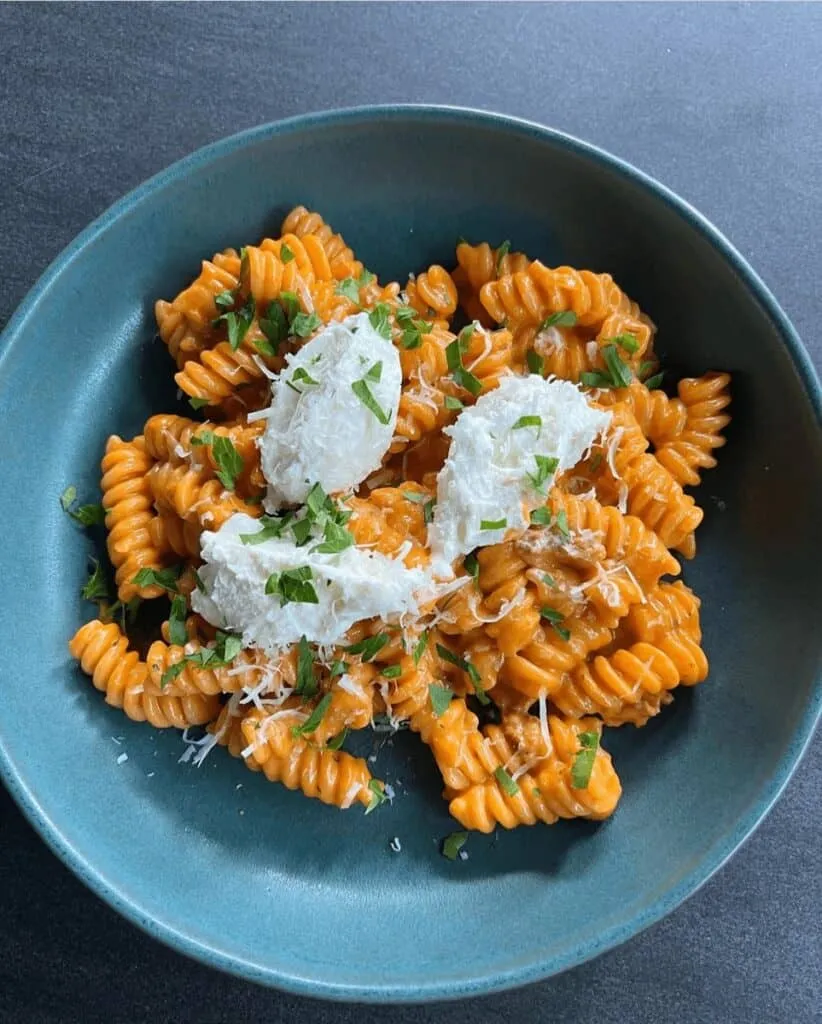 Creamy rotini pasta alla vodka with ricotta, garnished with fresh herbs in a blue bowl on a dark surface.