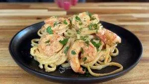 Creamy shrimp pasta garnished with fresh parsley on a black plate, set on a wooden table.