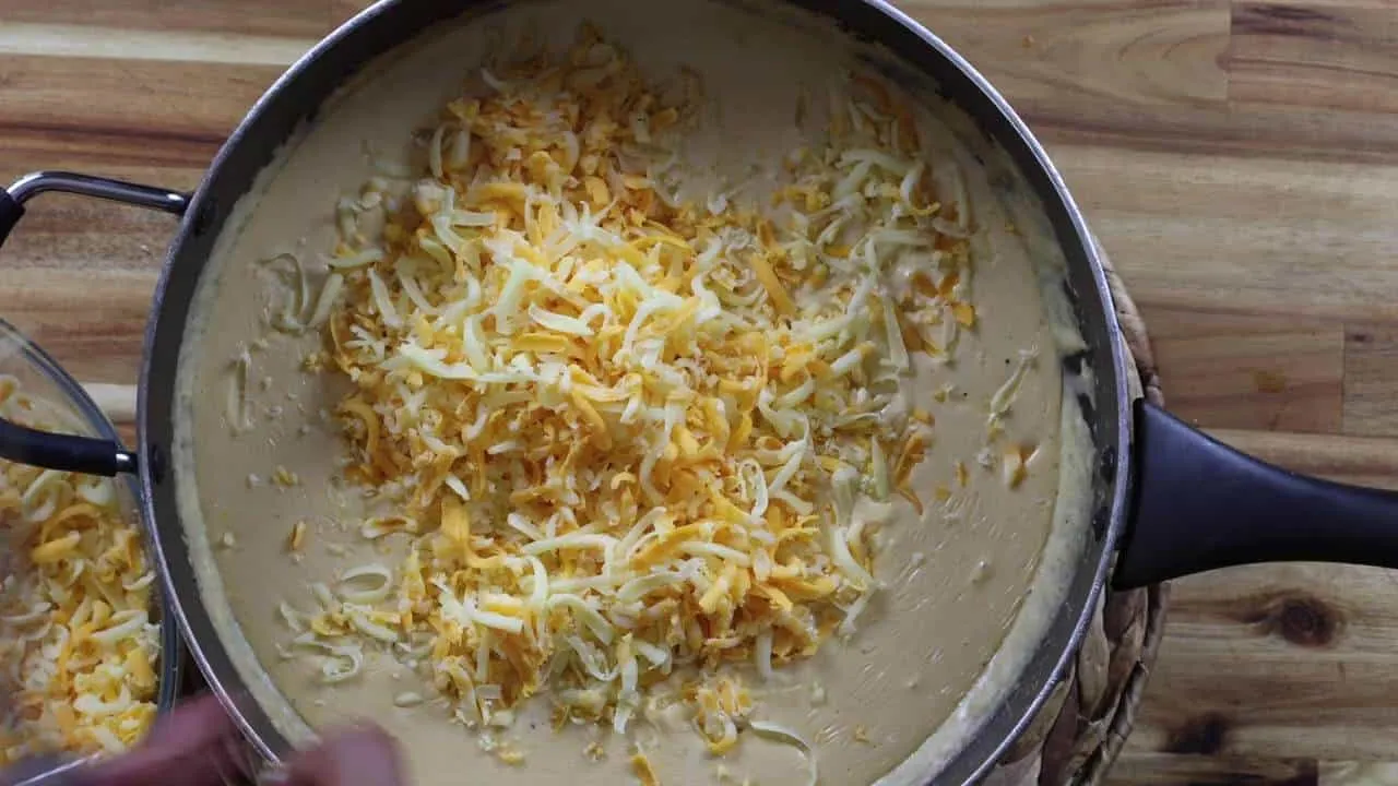 Pan of creamy cheese sauce topped with shredded cheddar and mozzarella on a wooden table.