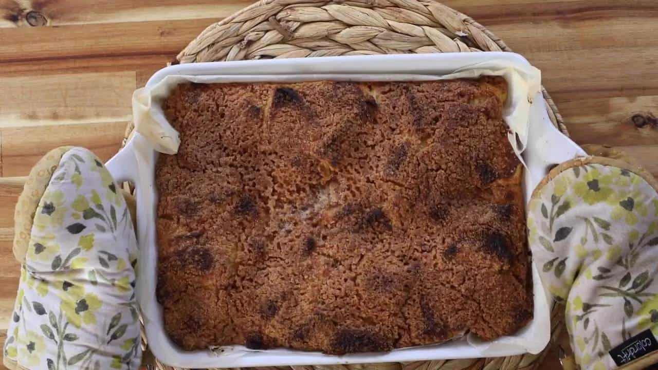 Freshly baked apple crumble in a white dish on a wooden table, with oven mitts holding it.