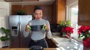 Woman in kitchen holding pot with oven mitts, surrounded by houseplants and holiday flowers.