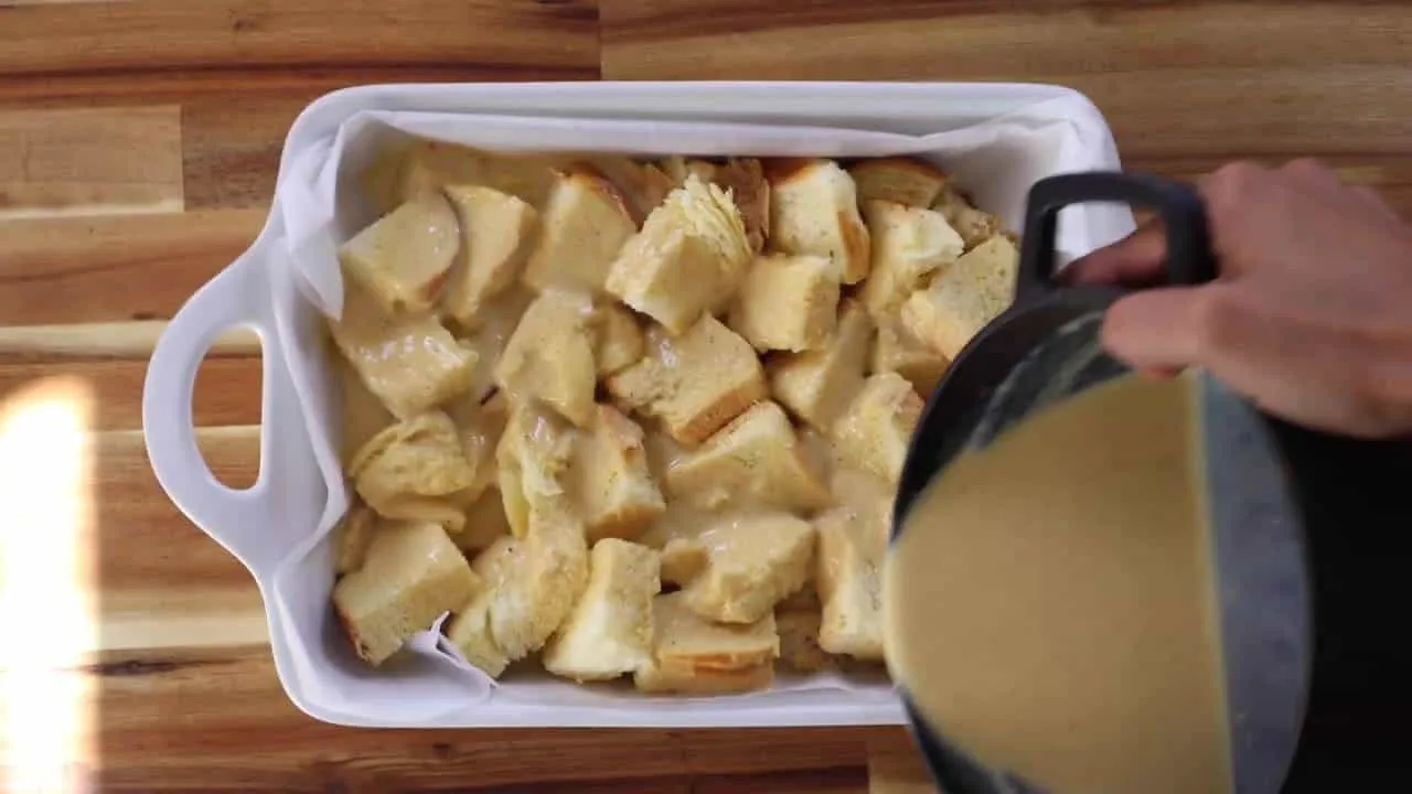 Pouring custard over bread cubes for homemade bread pudding in a white dish, ready for baking.