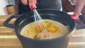 Whisking eggs into batter in a black pot on a wooden countertop, cooking preparation.