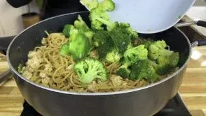 Broccoli being added to pasta with tofu in a pan on a wooden countertop.
