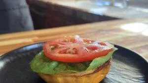 Open-faced sandwich with fresh tomato and lettuce on a toasted bun, placed on a black plate, in warm kitchen lighting.
