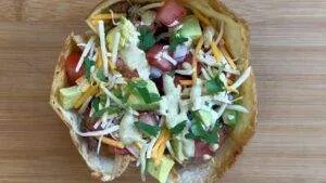Taco salad in a crispy shell topped with avocado, cheese, tomatoes, cilantro, and creamy dressing on a wooden board.