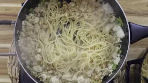 Spaghetti with tofu and green onions being cooked in a black pan on a wooden surface.
