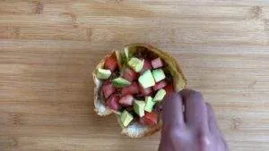 Taco salad with avocado and tomato pieces on a wooden board, hand arranging toppings for a fresh meal.