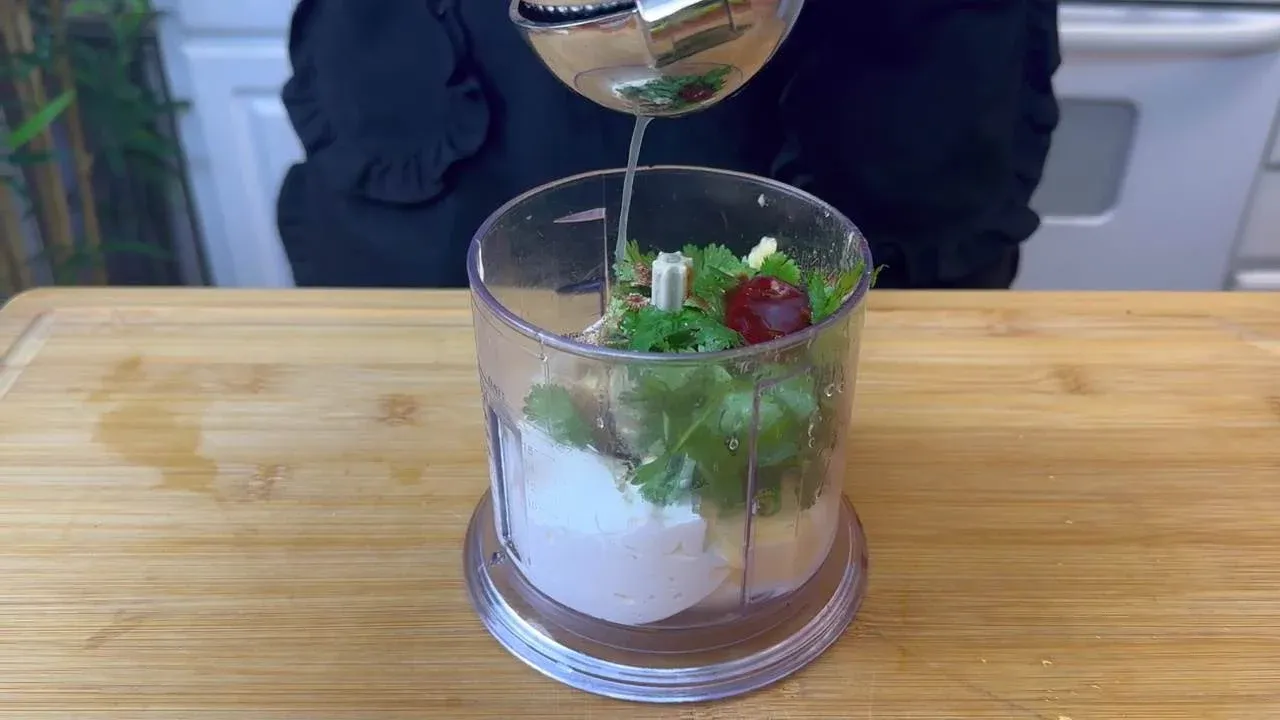 Pouring oil into a blender with herbs and spices, preparing fresh dressing on a wooden countertop.