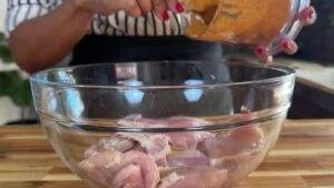 Adding marinade to raw chicken in a glass bowl on a kitchen counter.