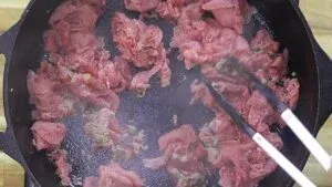 Cooking thinly sliced shaved beef in a cast iron skillet with tongs.