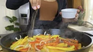 Person seasoning sliced bell peppers and onions in a hot skillet, creating a delicious sauté in a kitchen.