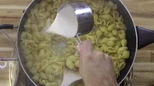 Pouring heavy cream into a pot of cooked shell pasta on a wooden countertop.