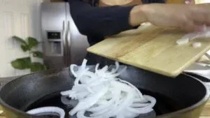 Person adding sliced onions to a frying pan from a wooden board in a kitchen.