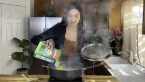 Woman cooking pasta in a kitchen, pouring from a box into a steaming pot on a stove.