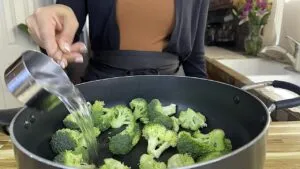 Person adding water to broccoli in a pan, preparing for steaming in a kitchen setting.