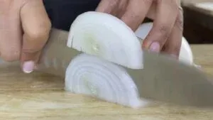 Close-up of hands slicing a white onion on a wooden cutting board with a knife.