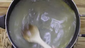 Wooden spoon stirring bubbling bone broth in a pot on a wooden board. Cooking preparation in progress.