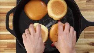 Toasting burger buns in a skillet with hands turning them gently.