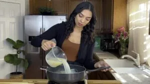 Woman pouring chicken broth into a pan in kitchen, preparing a recipe with fresh ingredients.