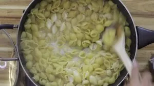 Cooking shell pasta in a pot with heavy cream, stirred with a wooden spoon on a wooden countertop.