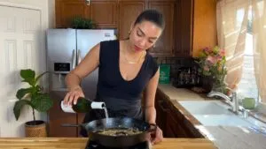Woman cooking in kitchen, pours wine into skillet, surrounded by home decor and sunlight filtering through curtains.