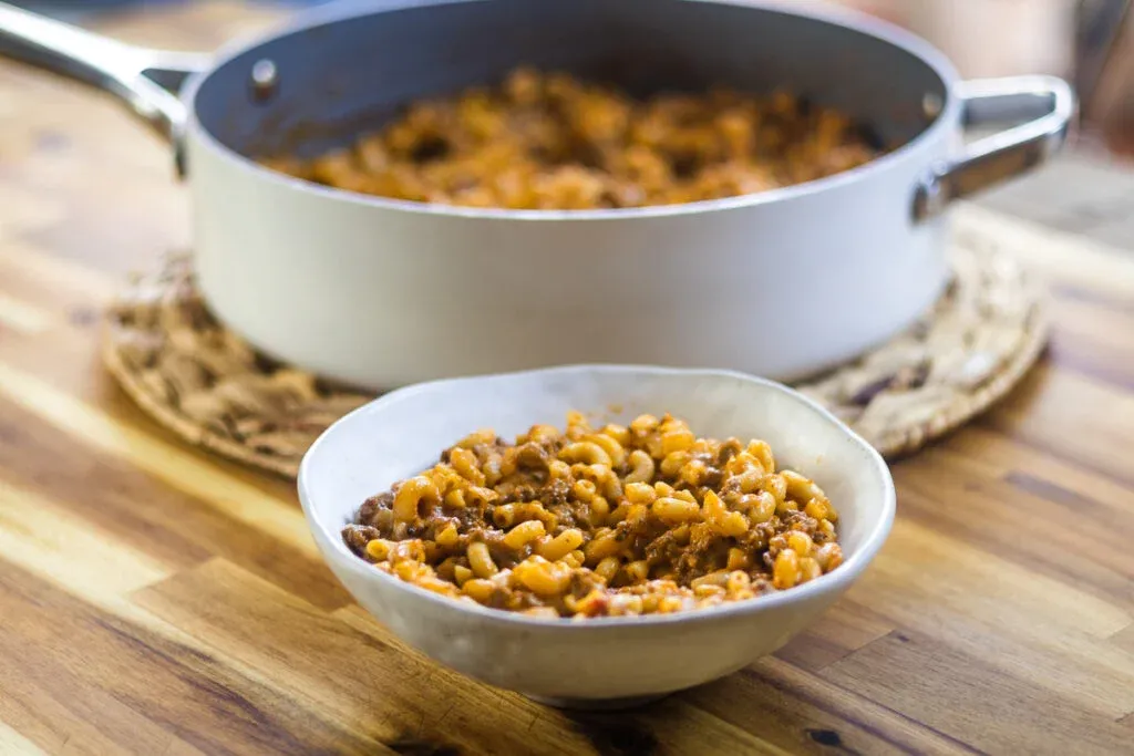 Bowl of savory macaroni beef skillet dish on a wooden table with cooking pot in background.