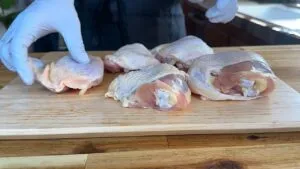 Person preparing raw chicken thighs on a wooden cutting board, wearing gloves.
