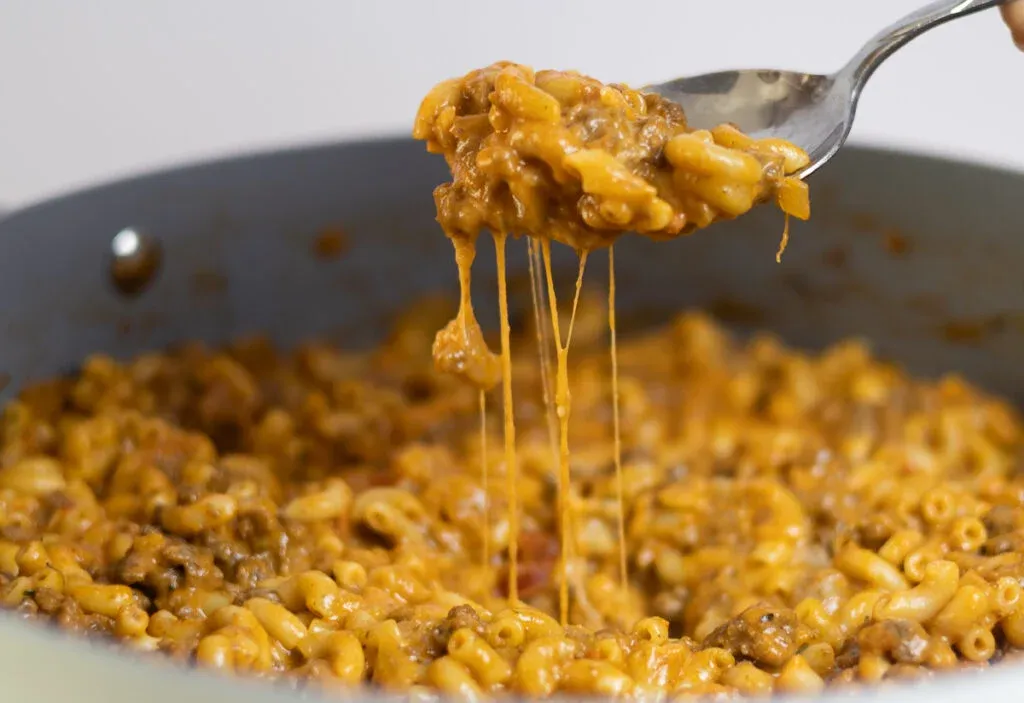 Cheesy beef macaroni in a skillet, with a spoon lifting a gooey, delicious portion.