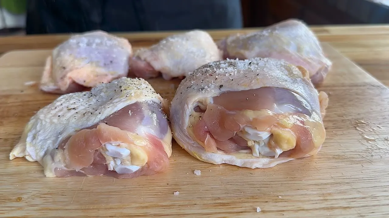 Raw chicken thighs seasoned with salt and pepper on a wooden cutting board.