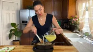 Woman cooking in kitchen, pouring chicken broth into skillet while stirring with wooden spoon.