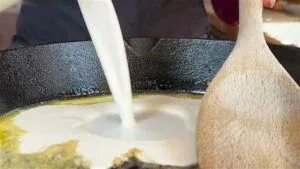 Pouring cream into a frying pan with a wooden spoon, cooking preparation close-up.