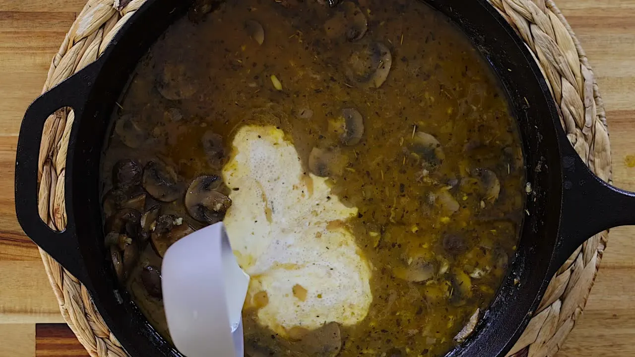 Adding cream to mushroom gravy in a black pot on a wooden table. Fresh and flavorful ingredients.