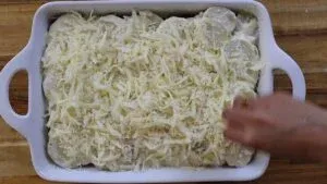 Person preparing cheesy gratin dish with shredded cheese in a baking dish on a wooden table.