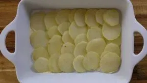 Sliced potatoes layered in a white baking dish on a wooden table, ready for cooking.