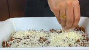 Sprinkling cheese over a beef dish in a baking tray, preparing a savory meal.
