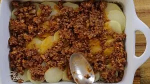Layered potato and meat casserole in white dish being prepared with a spoon.