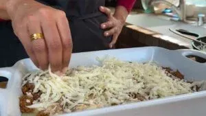 Person sprinkling shredded cheese over a casserole dish in a kitchen setting.
