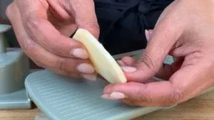 Hands holding a thin slice of potato over a mandoline slicer. Cooking preparation process.