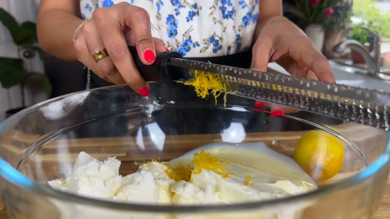 Grating lemon zest into cream cheese mixture in a glass bowl for a fresh dessert recipe.