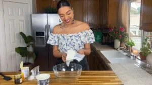 Woman preparing ingredients in a bright kitchen, holding cheese by a bowl on a wooden counter with flowers nearby.
