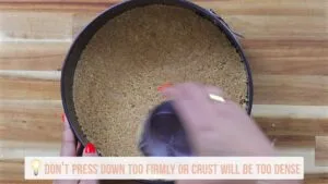 Person pressing graham cracker crust into a round pan with a glass on a wooden surface.