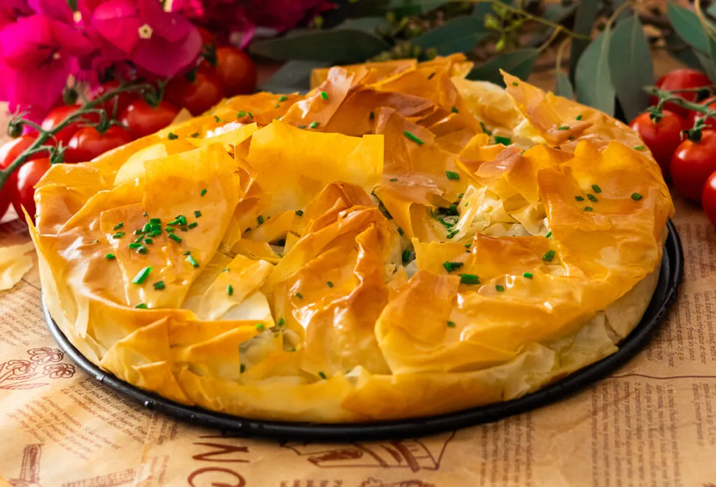 Crispy Mediterranean Spinach Pie with Puff Pasty garnished with chives, surrounded by fresh tomatoes and vibrant pink flowers.