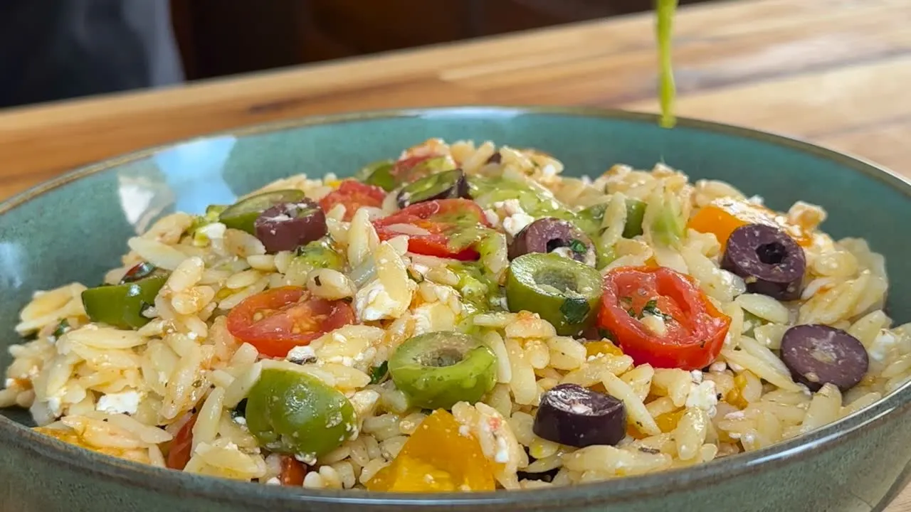 Dressing being drizzled on Colorful orzo salad with cherry tomatoes, olives, feta, and herbs in a green bowl on a wooden table.