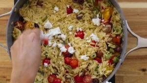 Colorful Mediterranean orzo with cherry tomatoes, olives, and feta being stirred in a pan.